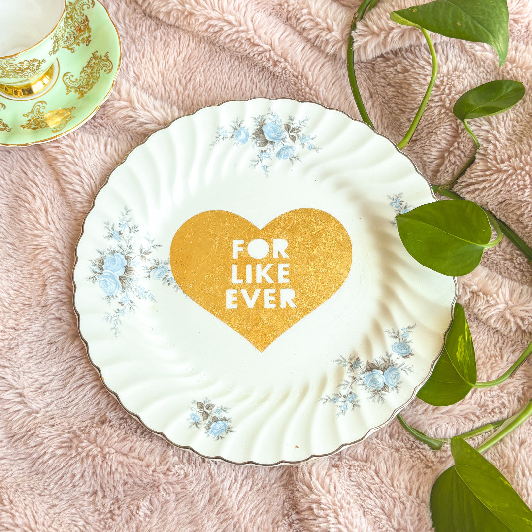 Decorative plate with a heart and 'FOR LIKE EVER' text on a pink surface with green leaves.