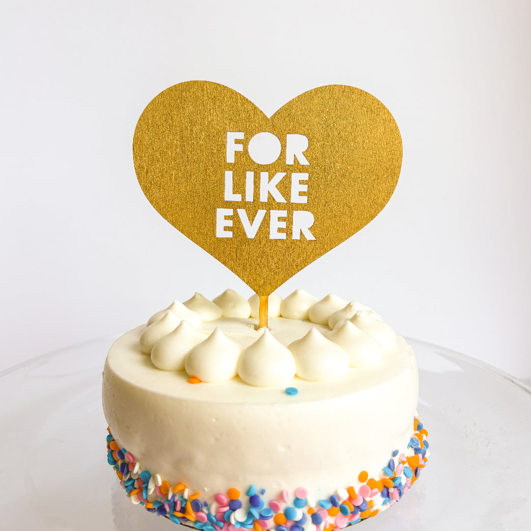 Small white cake with colorful sprinkles and a gold heart topper reading 'FOR LIKE EVER' on a white background.