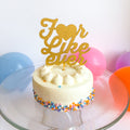 Small white cake with colorful sprinkles on a glass plate, topped with a 'I love you forever' gold cake topper, with balloons in the background.