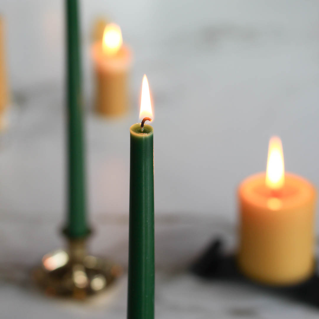 A lit green taper candle standing in a brass candlestick. There are other candles in the background, including two unlit green taper candles and two lit tea lights.