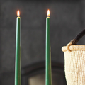 Two lit green taper candles standing upright in a candlestick. In the background, there is a wicker basket.