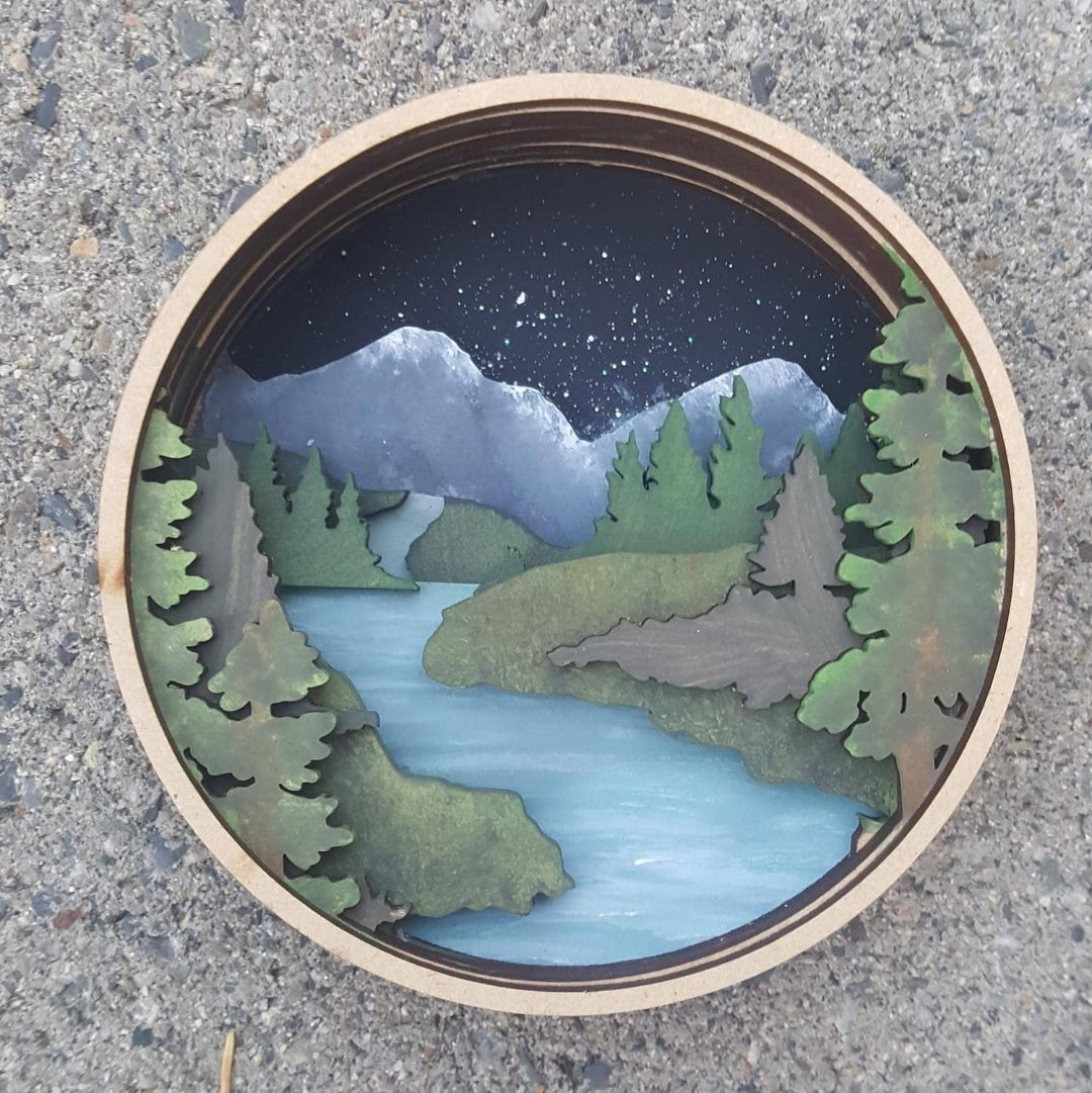 Wooden cutout of a scenic landscape with trees, mountains, and a lake on a concrete surface.
