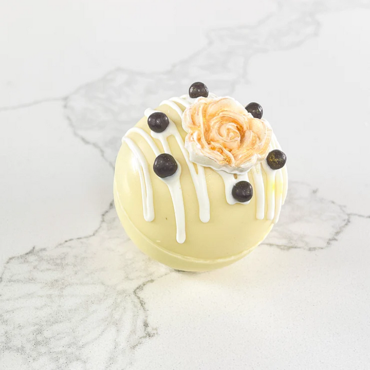 A white chocolate bath bomb decorated with a peach-colored rose and chocolate chips. The bath bomb is resting on a white marble surface.