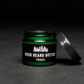 A dark green glass jar with a black lid. The jar is filled with beard butter and has a black label with the text "BUSH BEARD BUTTER FRESH" on it. The lid is slightly open and the jar is resting on a black surface.