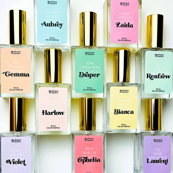 A collection of ten perfume bottles arranged in a grid. Each bottle has a gold cap and a transparent body, revealing the colored liquid inside. Each bottle has a label with the perfume's name and a list of its fragrance notes. The names include Aubrey, Zaida, Gemma, Draper, Harlow, Bianca, Renfrew, Ophelia, Laurent, and Violet.