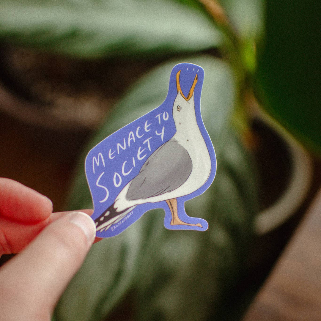 Hand holding a sticker with a bird design and text against a green leafy background