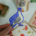 A hand holds a die-cut sticker featuring a stylized bird with the text 'MENACE TO SOCIETY' in white lettering on a blue background. The sticker has a gray body, white head, orange beak and legs, and is photographed against a softly blurred indoor background.