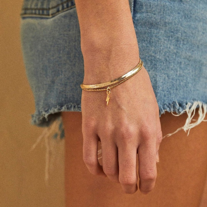 A set of two gold bracelets, one layered over the other, with a lightning bolt charm hanging from the bottom. The bracelets are displayed on a pink card with the brand name 
