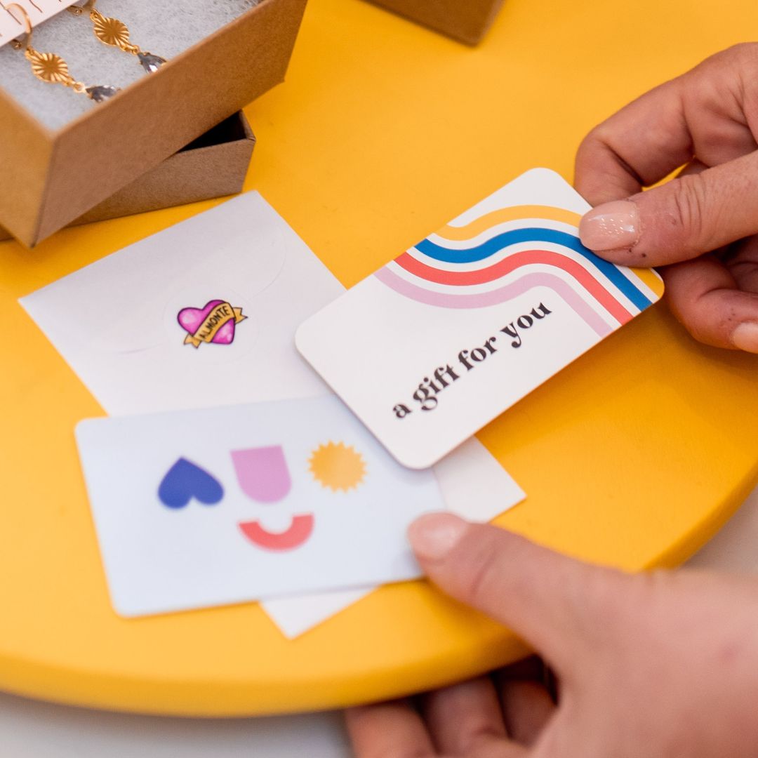 A person's hands holding a white gift card that reads 'a Gift for you' with a colorful rainbow design, next to other gift cards and a jewelry box on a bright yellow surface.