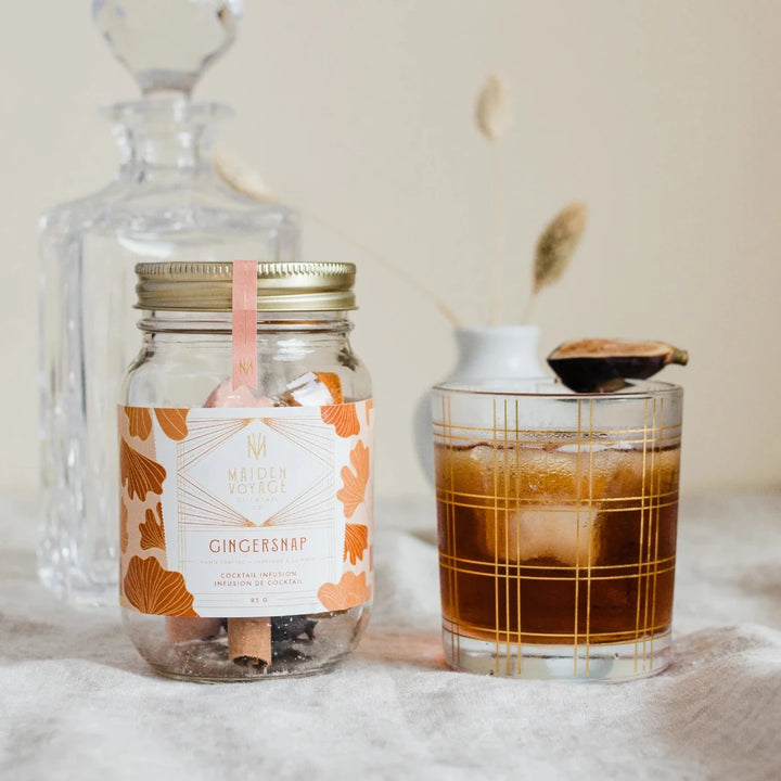 A glass jar filled with dried orange slices, cloves, and other spices. The jar has a white label with the Maiden Voyage logo and the product name 