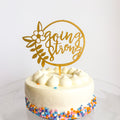 Small round cake with white frosting and colorful sprinkles, topped with a gold 'going strong' cake topper.