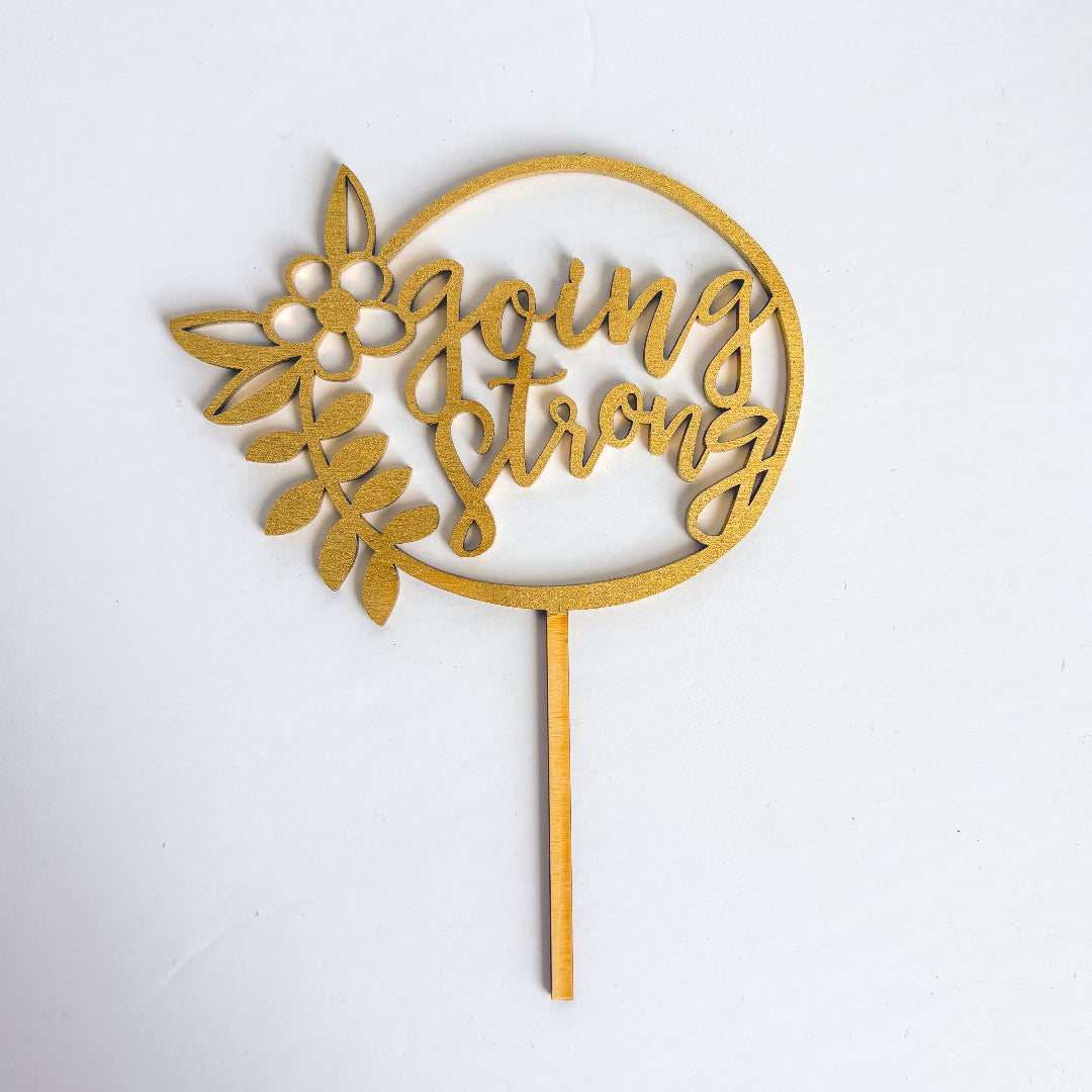 Gold cake topper with 'going strong' text on a white background