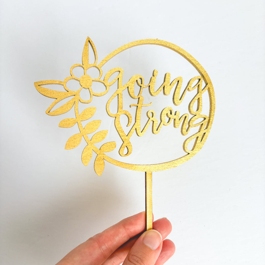 Gold decorative pick with 'going strong' text held by a hand against a white background