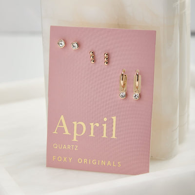 A set of three pairs of quartz earrings displayed on a pink card. The set includes a pair of stud earrings, a pair of bar earrings with small quartz beads, and a pair of hoop earrings with dangling quartz stones. The card has the month 