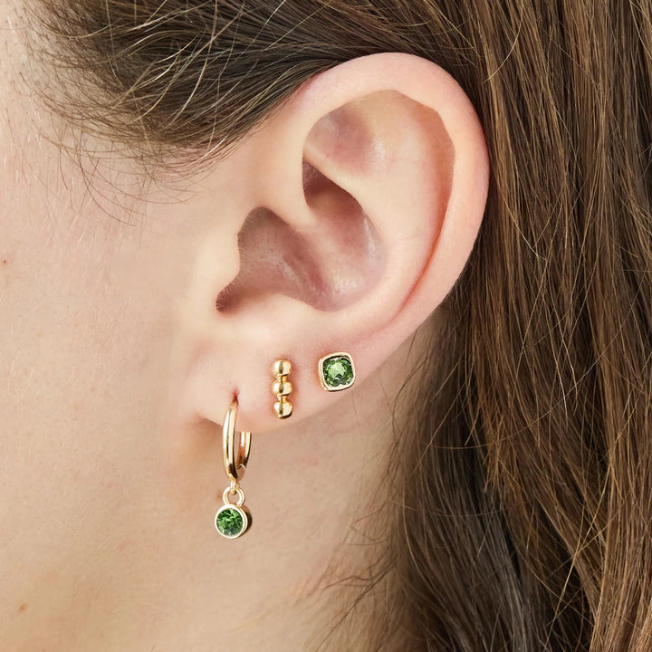 A set of three pairs of peridot earrings displayed on a green card. The set includes a pair of stud earrings, a pair of bar earrings with small peridot beads, and a pair of hoop earrings with dangling peridot stones. The card has the month 
