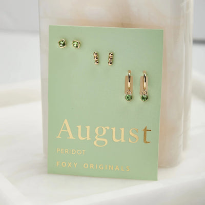 A set of three pairs of peridot earrings displayed on a green card. The set includes a pair of stud earrings, a pair of bar earrings with small peridot beads, and a pair of hoop earrings with dangling peridot stones. The card has the month 