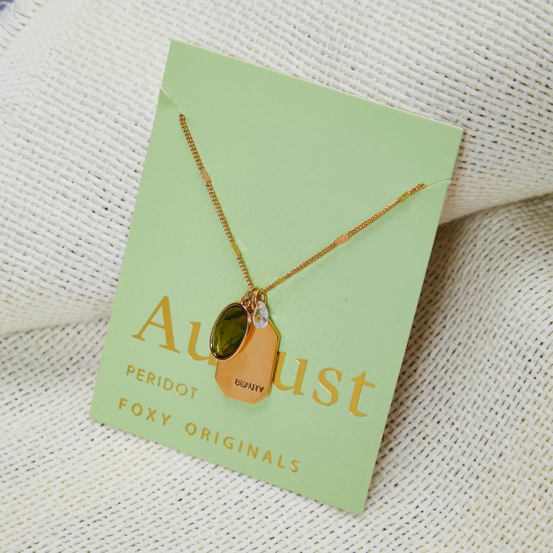 A gold necklace with a peridot stone pendant. The pendant also has the word "BEAUTY" engraved on it. The necklace is displayed on a green card with the month "August" and the brand name "Foxy Originals."