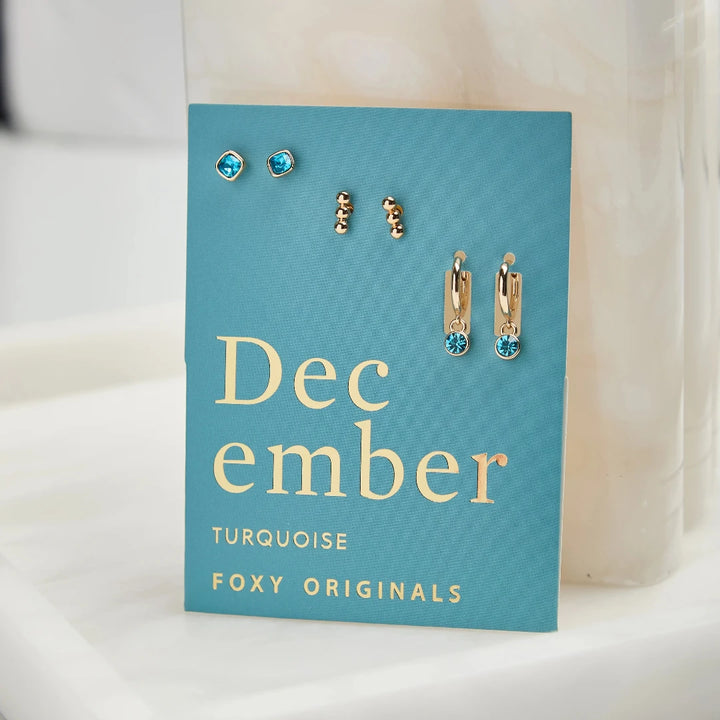 A set of three pairs of turquoise earrings displayed on a blue card. The set includes a pair of stud earrings, a pair of bar earrings with small turquoise beads, and a pair of hoop earrings with dangling turquoise stones. The card has the month 