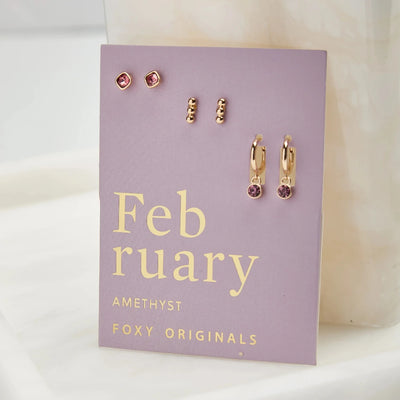A set of three pairs of amethyst earrings displayed on a purple card. The set includes a pair of stud earrings, a pair of bar earrings with small amethyst beads, and a pair of hoop earrings with dangling amethyst stones. The card has the month 