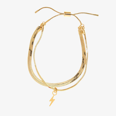 A set of two gold bracelets, one layered over the other, with a lightning bolt charm hanging from the bottom. The bracelets are displayed on a pink card with the brand name 