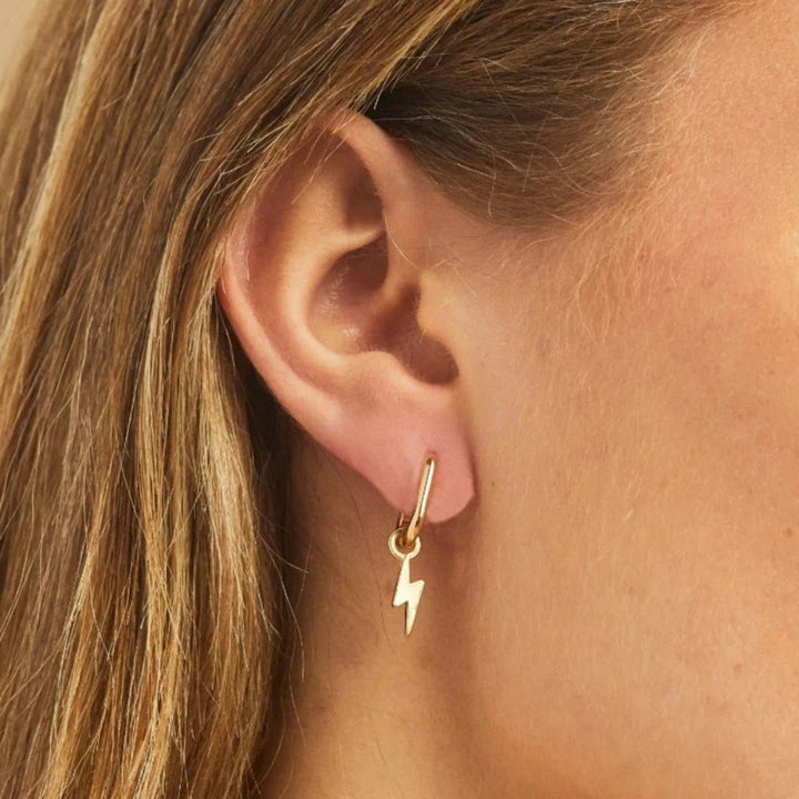 A pair of gold earrings with lightning bolt charms on a pink background. The earrings are displayed on a card with the brand name 