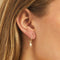 A pair of gold earrings with lightning bolt charms on a pink background. The earrings are displayed on a card with the brand name 