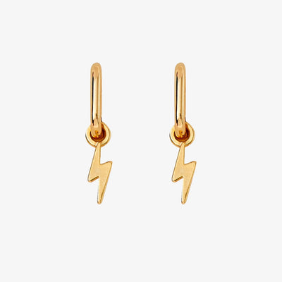 A pair of gold earrings with lightning bolt charms on a pink background. The earrings are displayed on a card with the brand name 