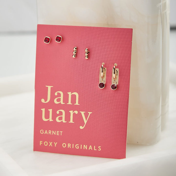 A set of three pairs of garnet earrings displayed on a red card. The set includes a pair of stud earrings, a pair of bar earrings with small garnet beads, and a pair of hoop earrings with dangling garnet stones. The card has the month 