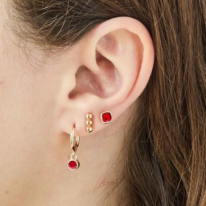 A set of three pairs of ruby earrings displayed on a red card. The set includes a pair of stud earrings, a pair of bar earrings with small ruby beads, and a pair of hoop earrings with dangling ruby stones. The card has the month 