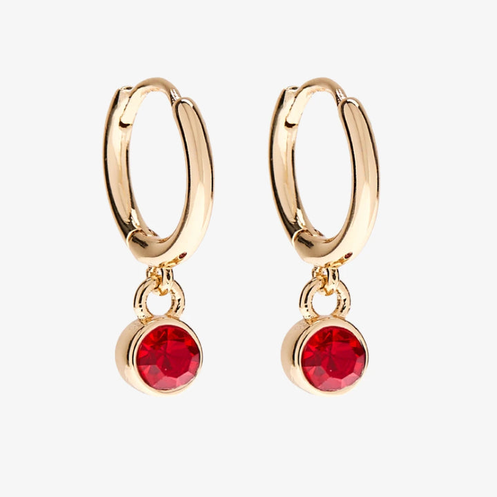 A set of three pairs of ruby earrings displayed on a red card. The set includes a pair of stud earrings, a pair of bar earrings with small ruby beads, and a pair of hoop earrings with dangling ruby stones. The card has the month 