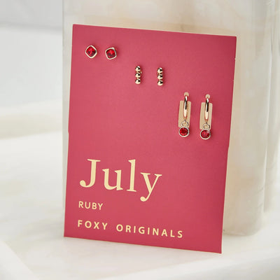 A set of three pairs of ruby earrings displayed on a red card. The set includes a pair of stud earrings, a pair of bar earrings with small ruby beads, and a pair of hoop earrings with dangling ruby stones. The card has the month 
