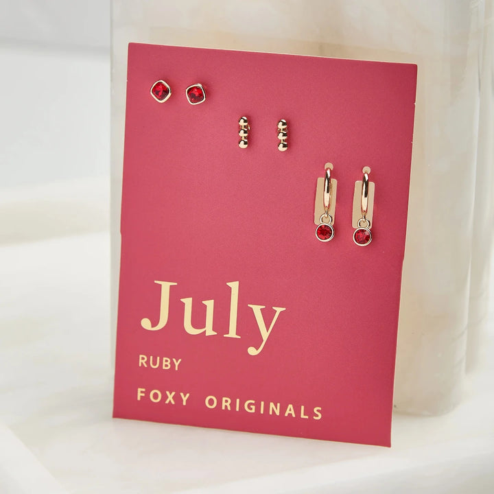 A set of three pairs of ruby earrings displayed on a red card. The set includes a pair of stud earrings, a pair of bar earrings with small ruby beads, and a pair of hoop earrings with dangling ruby stones. The card has the month 