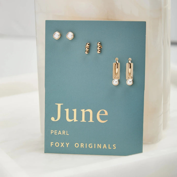 A set of three pairs of pearl earrings displayed on a blue card. The set includes a pair of stud earrings, a pair of bar earrings with small pearl beads, and a pair of hoop earrings with dangling pearl stones. The card has the month 