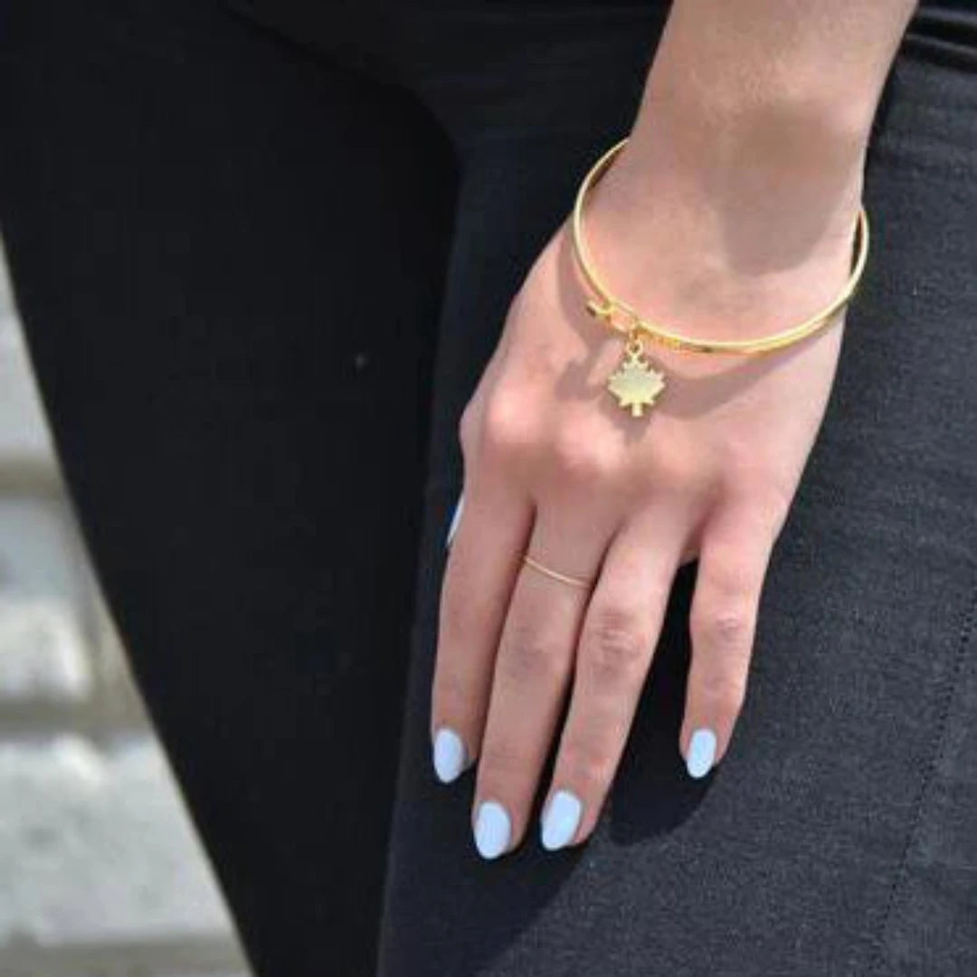 A gold bangle bracelet with a maple leaf charm. The bracelet is displayed on a card with a photo of a Canadian cabin in the woods and the hashtag #CANADA150. The card also includes the information "MADE IN CANADA" and the brand name "Foxy Originals."