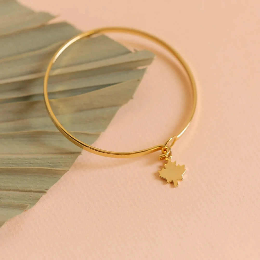 A gold bangle bracelet with a maple leaf charm. The bracelet is displayed on a card with a photo of a Canadian cabin in the woods and the hashtag #CANADA150. The card also includes the information "MADE IN CANADA" and the brand name "Foxy Originals."