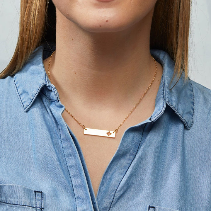 A gold necklace with a bar pendant. The bar has a cut-out maple leaf design.