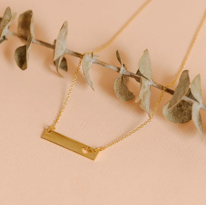 A gold necklace with a bar pendant. The bar has a cut-out maple leaf design.