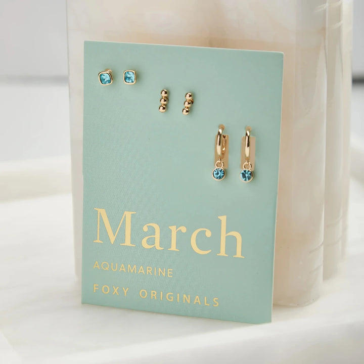 A set of three pairs of aquamarine earrings displayed on a light blue card. The set includes a pair of stud earrings, a pair of bar earrings with small aquamarine beads, and a pair of hoop earrings with dangling aquamarine stones. The card has the month 