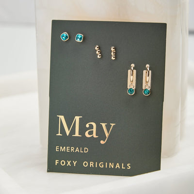 A set of three pairs of emerald earrings displayed on a green card. The set includes a pair of stud earrings, a pair of bar earrings with small emerald beads, and a pair of hoop earrings with dangling emerald stones. The card has the month 