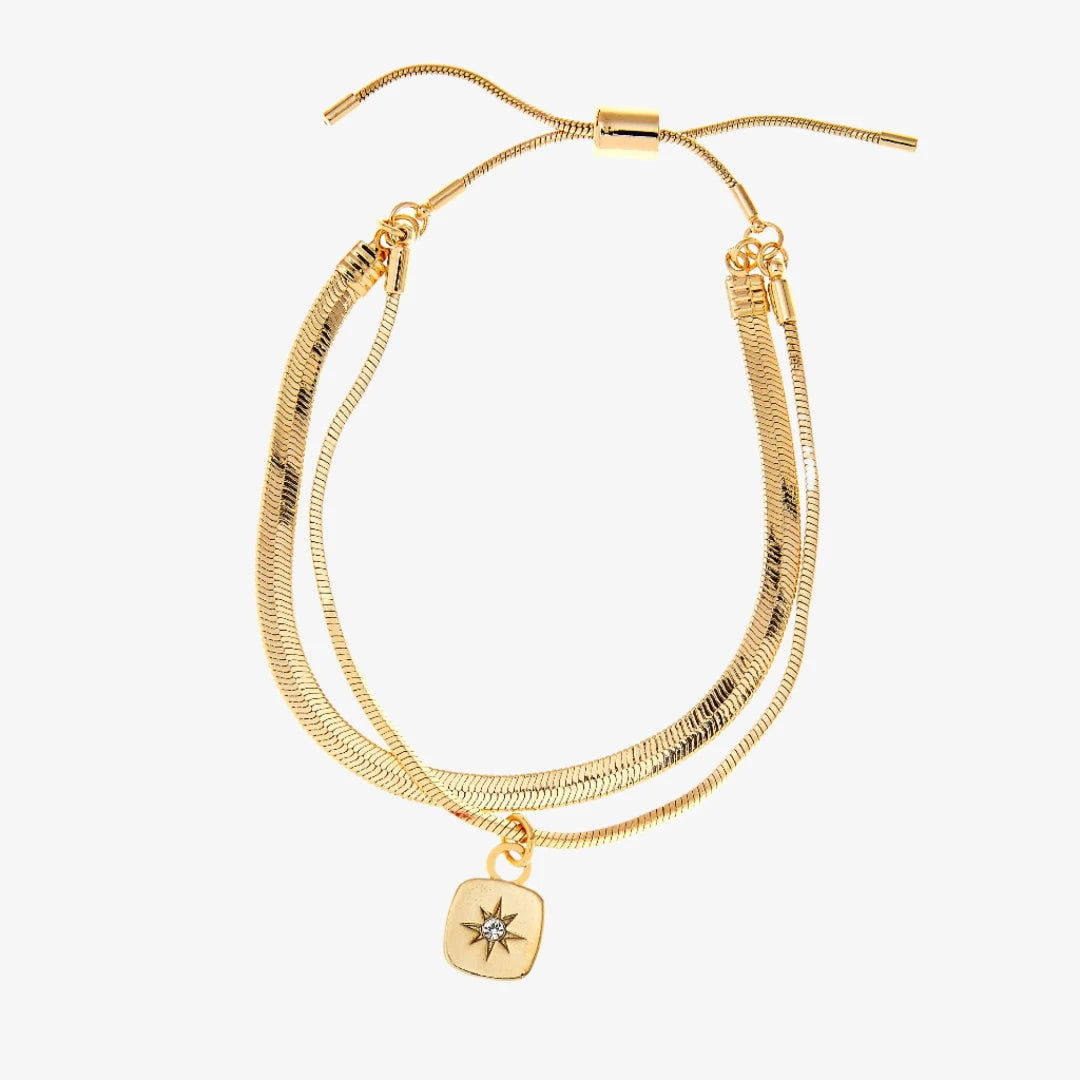 A set of two gold bracelets, one layered over the other, with a star charm hanging from the bottom. The bracelets are displayed on a pink card with the brand name "Foxy Originals."