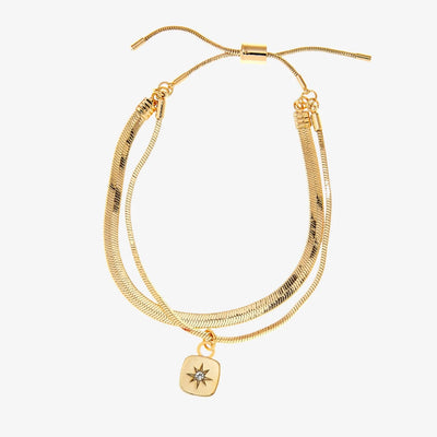 A set of two gold bracelets, one layered over the other, with a star charm hanging from the bottom. The bracelets are displayed on a pink card with the brand name 