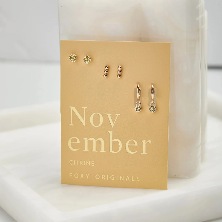 A set of three pairs of citrine earrings displayed on a yellow card. The set includes a pair of stud earrings, a pair of bar earrings with small citrine beads, and a pair of hoop earrings with dangling citrine stones. The card has the month 