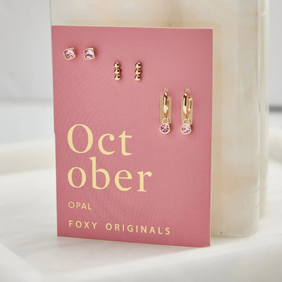 A set of three pairs of opal earrings displayed on a pink card. The set includes a pair of stud earrings, a pair of bar earrings with small opal beads, and a pair of hoop earrings with dangling opal stones. The card has the month 