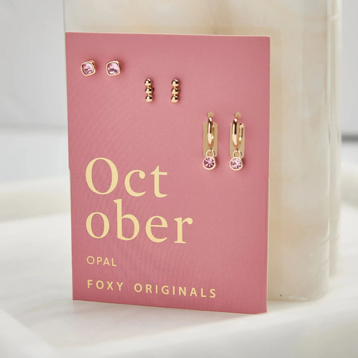 A set of three pairs of opal earrings displayed on a pink card. The set includes a pair of stud earrings, a pair of bar earrings with small opal beads, and a pair of hoop earrings with dangling opal stones. The card has the month 