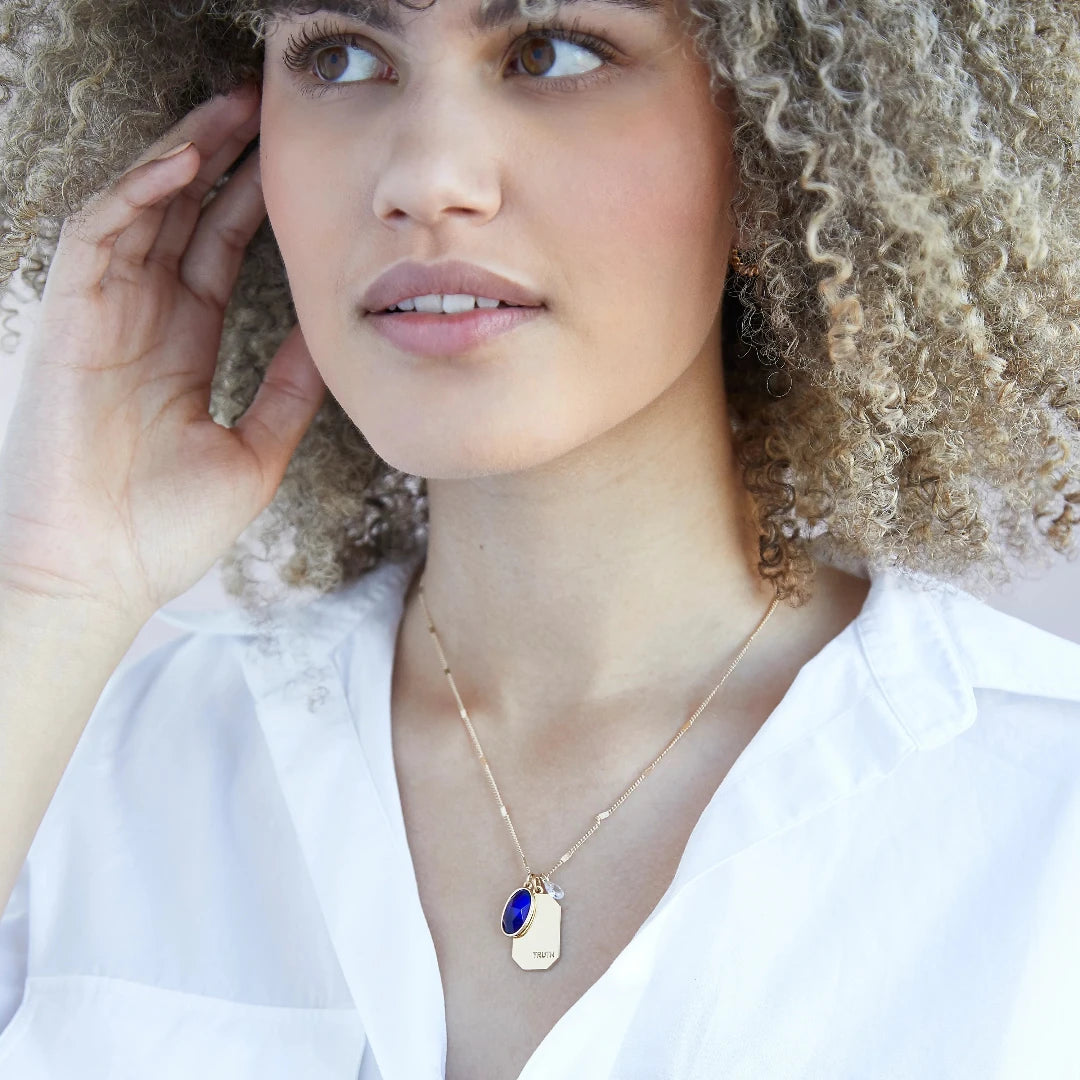 A gold necklace with a sapphire stone pendant. The pendant also has the word "TRUTH" engraved on it. The necklace is displayed on a blue card with the month "September" and the brand name "Foxy Originals."