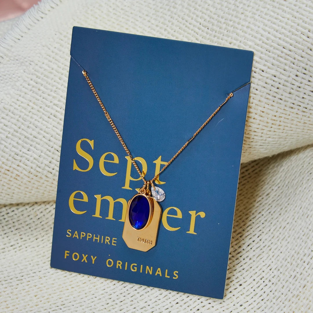 A gold necklace with a sapphire stone pendant. The pendant also has the word "TRUTH" engraved on it. The necklace is displayed on a blue card with the month "September" and the brand name "Foxy Originals."