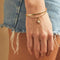 A set of two gold bracelets, one layered over the other, with a sun charm hanging from the bottom. The bracelets are displayed on a pink card with the brand name 