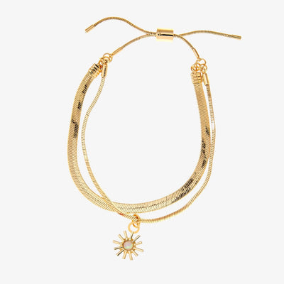 A set of two gold bracelets, one layered over the other, with a sun charm hanging from the bottom. The bracelets are displayed on a pink card with the brand name 