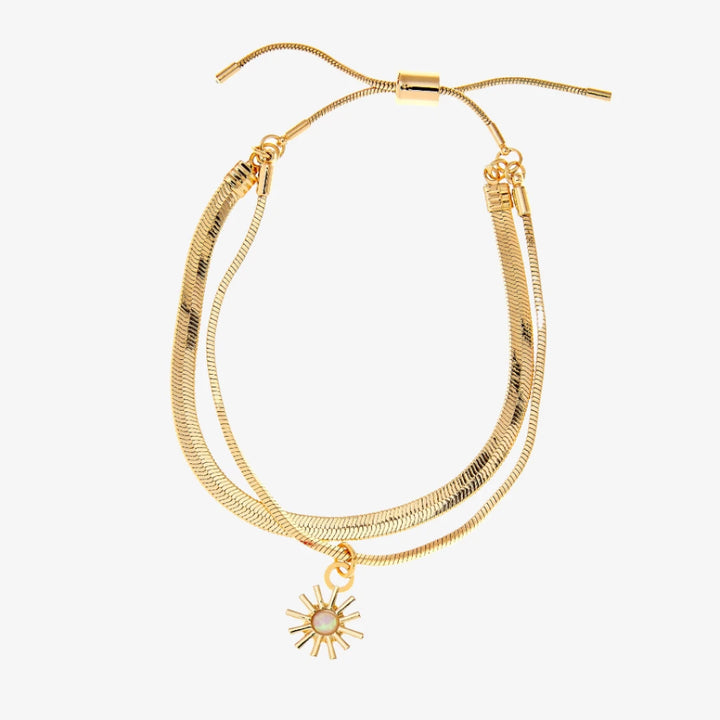 A set of two gold bracelets, one layered over the other, with a sun charm hanging from the bottom. The bracelets are displayed on a pink card with the brand name 