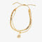 A set of two gold bracelets, one layered over the other, with a sun charm hanging from the bottom. The bracelets are displayed on a pink card with the brand name 